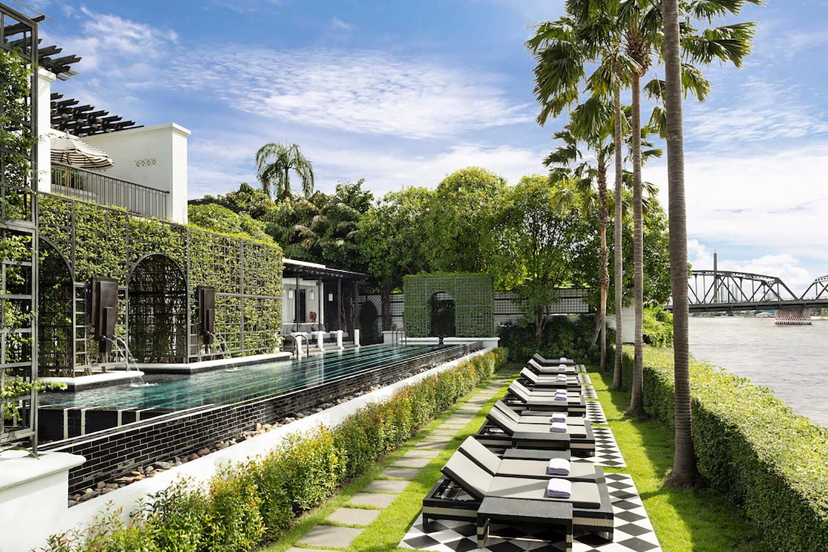 Outdoor pool area with lounge chairs, greenery, and a building in the background.