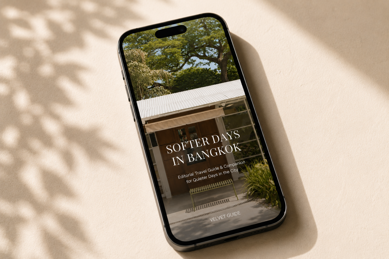 A black smartphone lying on a warm beige surface under dappled sunlight, displaying the cover of Softer Days in Bangkok travel guide